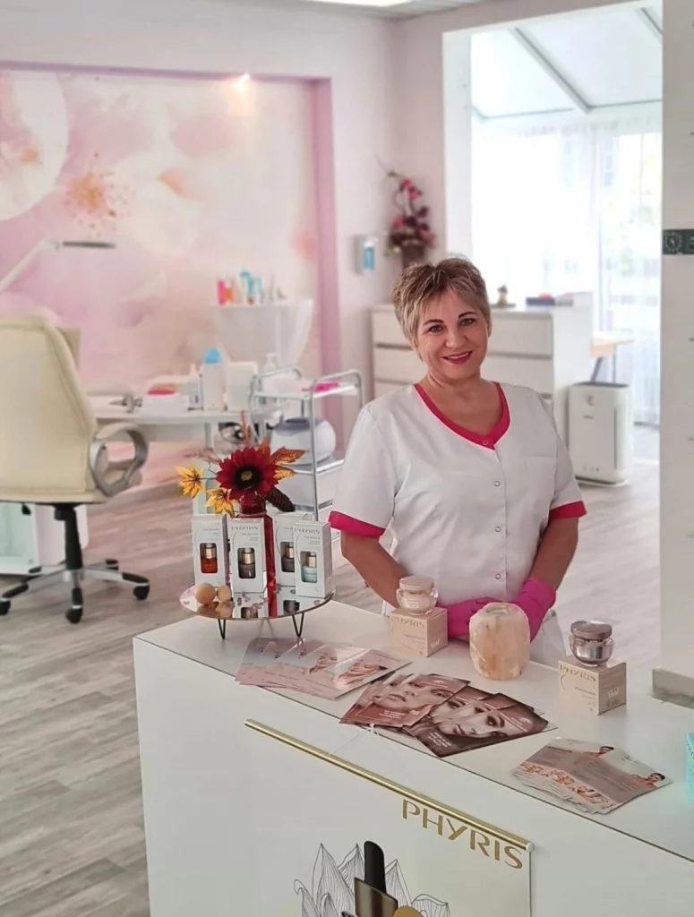Ein Innenraum eines Kosmetikstudios mit hellrosa Wänden, die mit floralen Designs geschmückt sind und einer Vielzahl von Beautyprodukten auf einem weißen Tresen. Elena steht hinter dem Tresen.